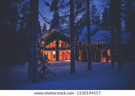 A night view of cozy wooden scandinavian cabin cottage chalet house covered in snow near ski resort in winter with the lights turn on, evening picture
