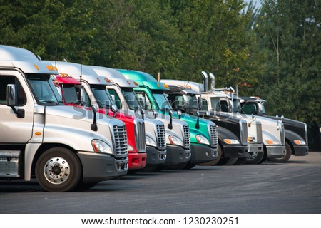 Big rigs semi trucks of different brands models and colors are lined up in parking lots truck stops rest areas filling vacant places to rest have lunch or wait for cargo following traffic schedule Royalty-Free Stock Photo #1230230251