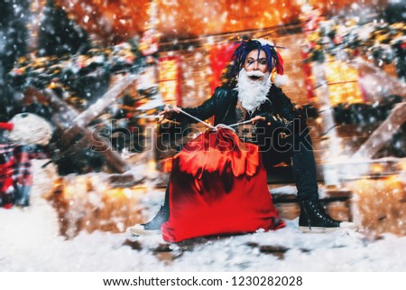 Сheerful punk Santa fools around near his house with a bag of gifts in his hands.