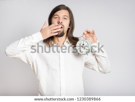 handsome long haired man is yawning with alarm clock, isolated on background