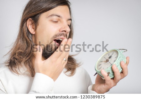 handsome long haired man is yawning with alarm clock, isolated on background