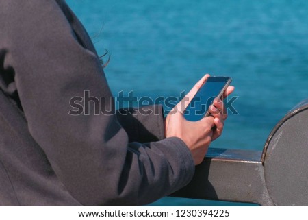 Brunette woman types a message on a mobile phone standing on the waterfront by the sea on a Sunny autumn day. Hands close-up.