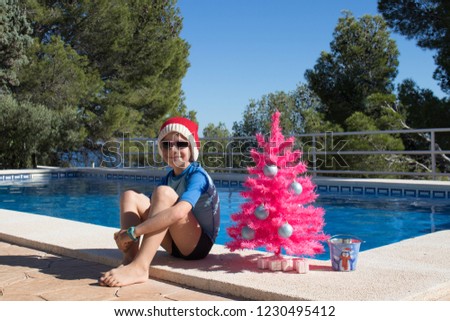 Happy Christmas Holidays. A cute child wearing a Santa hat by the swimming pool with a pink xmas tree. Merry Christmas Postcard Design with Copy Space. Christmas vacation in warm countries.
