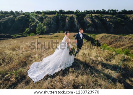 The young groom holds the hand of a beautiful bride on the background of the hills in sunny weather. Happy newlyweds are standing in a yellow field. Wedding photography. Family portrait.