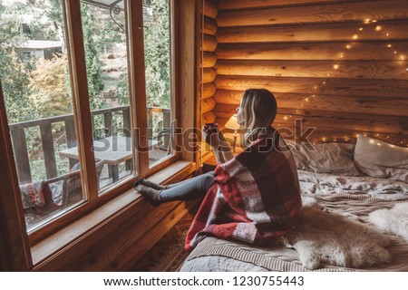Woman in warm blanket relaxing and drinking morning coffee on cozy bed in log cabin in winter Royalty-Free Stock Photo #1230755443