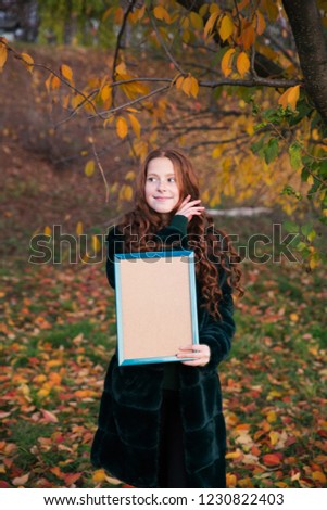 Red-hair girl with empty picture frame in the autumn garden full of colorful leaves.