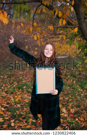 Red-hair girl with empty picture frame in the autumn garden full of colorful leaves.