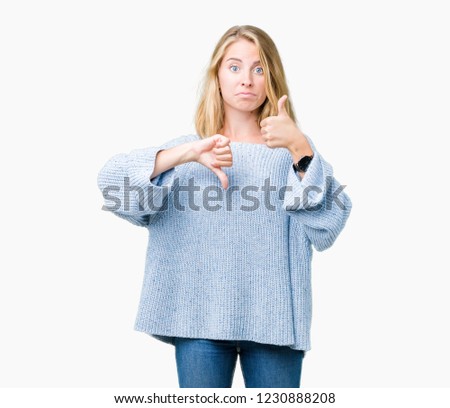 Beautiful young woman wearing blue sweater over isolated background Doing thumbs up and down, disagreement and agreement expression. Crazy conflict