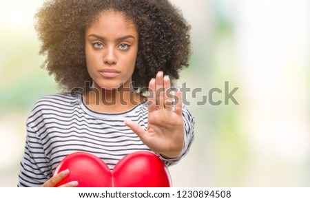 Young afro american woman holding red heart in love over isolated background with open hand doing stop sign with serious and confident expression, defense gesture