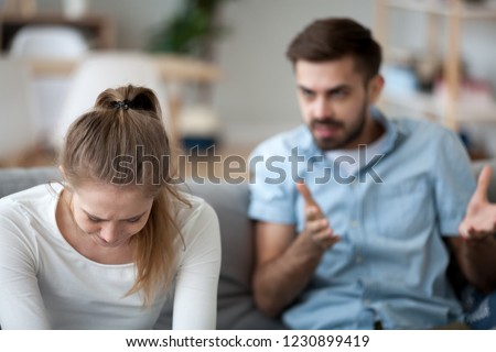 Upset wife sit on couch crying listening to furious husband yelling, unhappy couple have fight or disagreement at home, sad woman feel desperate and down with mad spouse scolding and lecturing Royalty-Free Stock Photo #1230899419