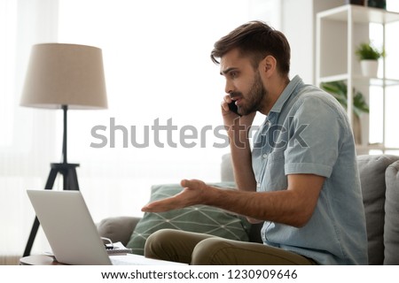 Confused male having computer problems talk on mobile with customer support, frustrated millennial man speak on smartphone managing computer malfunction working online from home Royalty-Free Stock Photo #1230909646