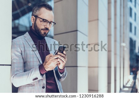 Curious bearded man standing next to the office building and feeling interested while looking at the screen of his device