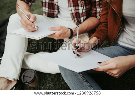Painting lesson. Young couple sitting on grass and holding notebooks. Close up of white sheets on knees of man and woman
