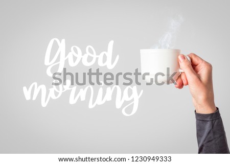 Female hand in clothes holding a white cup with hot coffee or tea on a light grey background. Added text Good morning. Breakfast concept with hot coffee or tea