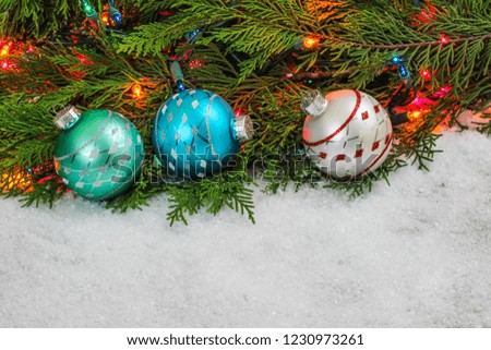 Christmas background with teal blue, red and silver ornaments, colorful string of lights and green Christmas tree garland border in snow; holiday background with white copy space 