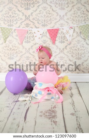 Cute happy blond baby girl in pink and yellow tutu and flower head band sitting on vintage wooden floor and background by smashed double tier polka dot decorated pink blue and white fondant iced cake