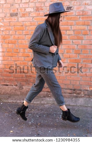 a stylish woman in a classic women's suit consisting of trousers and a jacket in a hood in the style of a gangster, posing for a photo against the background of a brick wall