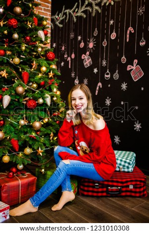 A seductive blonde in a red sweater sitting near a Christmas tree with gifts