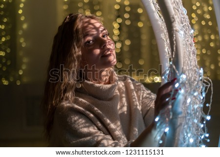 A beautiful blonde is sitting in a hanging chair on the background of Christmas garlands. A young woman in a white sweater on a background of Christmas lights.