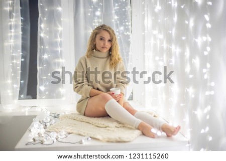 A beautiful blonde in a light sweater drinks a warming drink while sitting on a plaid against the backdrop of garlands.