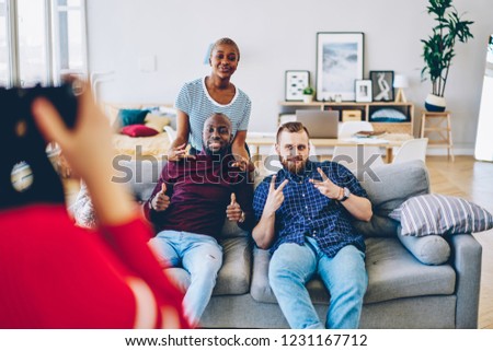 Multiracial smiling male and female friends spending time together at apartment posing for picture, young cheerful hipsters sitting on sofa at living room while woman taking photo on free time