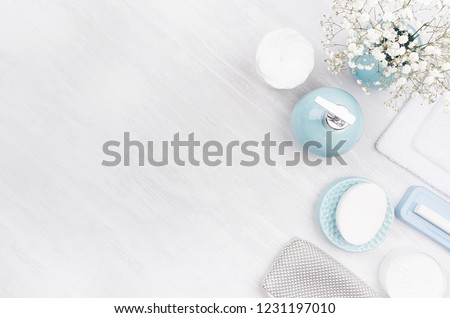 Flat lay of different cosmetics products and accessories of silver, pastel blue and white color with small flowers on white wooden background, copy space.