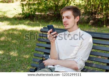 Young handsome hipster guy smoking vape while surfing the web on a smartphone using mobile internet. Smiling tudent male relaxing in the park while chatting online by a mobile phone.