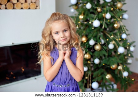 Happy little girl in purple luxury dress holds hands near face.With colorful lights and Christmas tree on background. Holidays, christmas, new year, x-mas concep.
