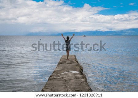 Young girl with her back to the camera, is standing on a long rock into the sea, cheering to blue sea, with blue sky and white clouds in background. 