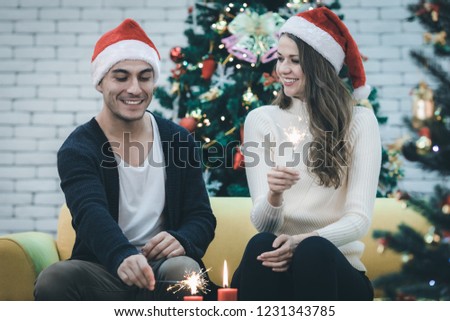 Smiling young good looking Caucasian couple light the candles and play mini firework, sitting on sofa together, in christmas time, concept for holiday season.