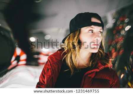 Trendy charming girl in red jacket wearing stylish cap with pensive face looking away. False glass is on background