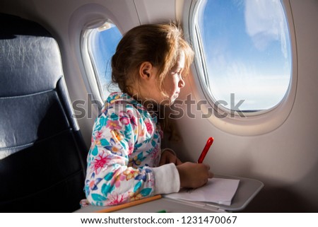 Child drawing picture with crayons in airplane. Little girl occupied while flying in aircraft. Travel with family and kids. Blue sky and sun outside the window