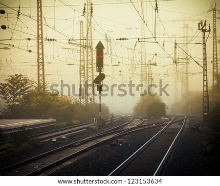 Moble photography lo-fi styled image of confusing urban railway tracks with ines and overhead cables and a red signal light at foggy dusk