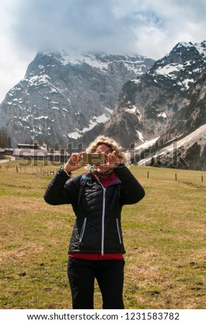 Young blonde woman takes a picture in front of mountain panorama with her smartphone