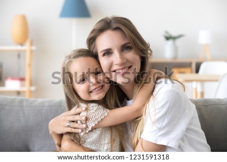 Happy loving young mother hugging little preschool daughter, smiling woman caring mom embracing kid girl sitting on couch at home, mommy and child looking at camera, warm family relations, portrait Royalty-Free Stock Photo #1231591318