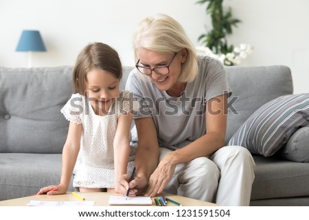 Smiling grandmother and little kid granddaughter drawing on paper with colored pencils together, caring granny teaching grandchild having fun playing at home, grandma and creative child activity