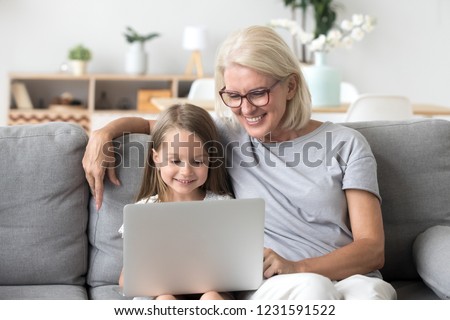 Smiling senior grandma and cute little kid granddaughter watching cartoons on laptop together, happy older grandmother playing game or shopping ordering buying online with child girl on computer