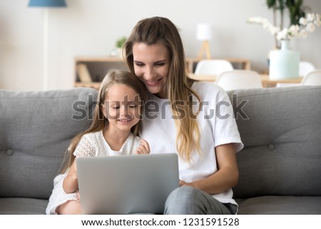 Smiling mother and kid daughter having fun shopping online sitting on sofa, happy mom baby sitter teaching little child girl use laptop application, watching cartoon or making video call on computer