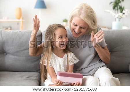 Excited grandchild happy to receive birthday present from loving old grandmother, smiling little kid holding gift box enjoys senior grandma surprise sitting on couch, granny congratulating child girl