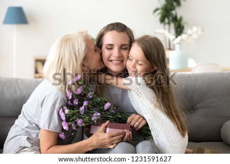 Little kid daughter and old mother kissing embracing young woman congratulating with birthday, senior grandmother and child girl presenting happy mom flowers gift box, 3 generation celebrate together