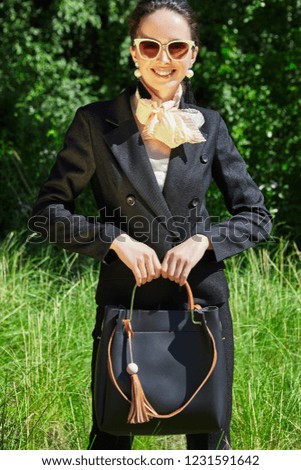 Three quarter portrait of a young lady in a black formal suit, a creamy neckerchief, sunglasses. The beautiful lady standing, posing in the park with a black handbag, smiling, looking at the camera.