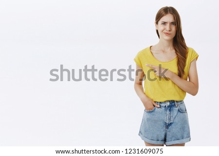 Girl points left smirking and frowning being doubtful with quality of product standing displeased and hesitant over gray background in yellow t-shirt expressing disappointment and dissatisfaction