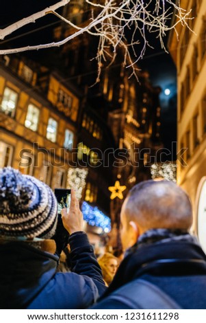 Adult male couple taking photo on smartphone of Notre-Dame Cathedral and maison Kammerzell decorated with Christmas lights and full moon seen in the blue clouds.