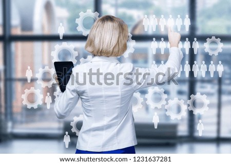 A businesswoman is operating with a digital system consisting of cogwheels and figures on the touchscreen at the office interior background. The concept is principle of interactive business.