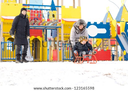Young family out toboganning in snow with mother pushing her young son on a colourful orange sled in front of a childrens playground