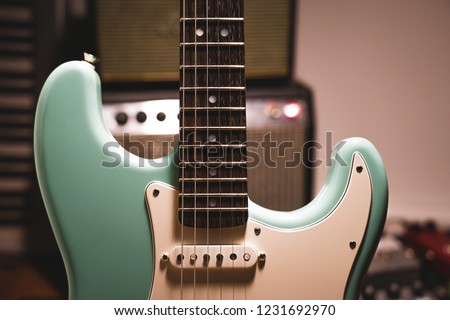 Closeup of seafoam green electric guitar with wooden wenge fretboard and mother of pearl inlays, white pickup and pickguard in front of blurry background with amps music studio