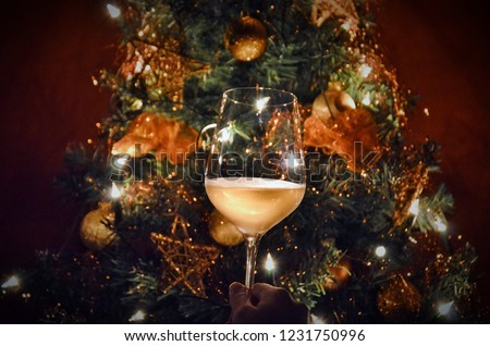 Woman hand proposing toast with a glass in front of the lighting xmas tree. The Christmas tree is decorated with beautiful ribbons and stars in golden color. 