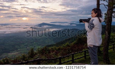 Woman tourist holding a DSLR camera looking at the beautiful nature landscape of sun fog mountain in the winter during sunrise on high viewpoint at Phu Ruea National Park, Loei province, Thailand