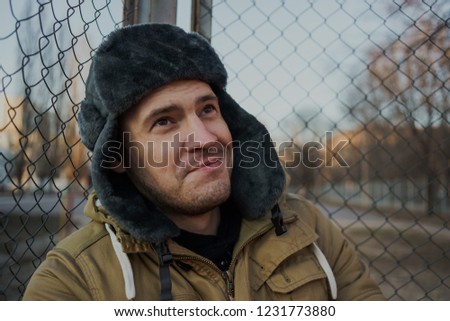 Happy handsome young man in cap with earflaps. The young man in the fur hat. a young guy standing on the street on a cloudy day. emotional portrait of a student. street style
