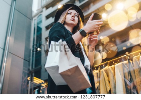Young blonde hipster girl in trendy outfit standing in front of show-window with paper shopping bags and mobile phone in hands.